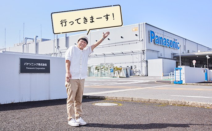 写真：ムラヨシマサユキさんがパナソニック株式会社の前に立ち「行ってきまーす！」と左手をあげている様子