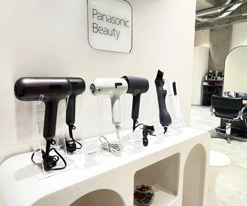Panasonic Beautyのドライヤーや美容家電が並ぶ展示スペースと、奥にカットチェアやミラーが配置された美容室の内観
