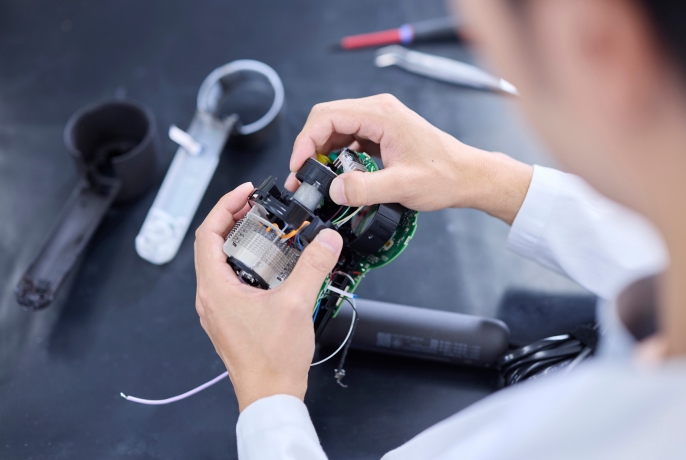 作業台の上で電子機器の内部部品を手に持ち、組み立てや検査を行っている様子