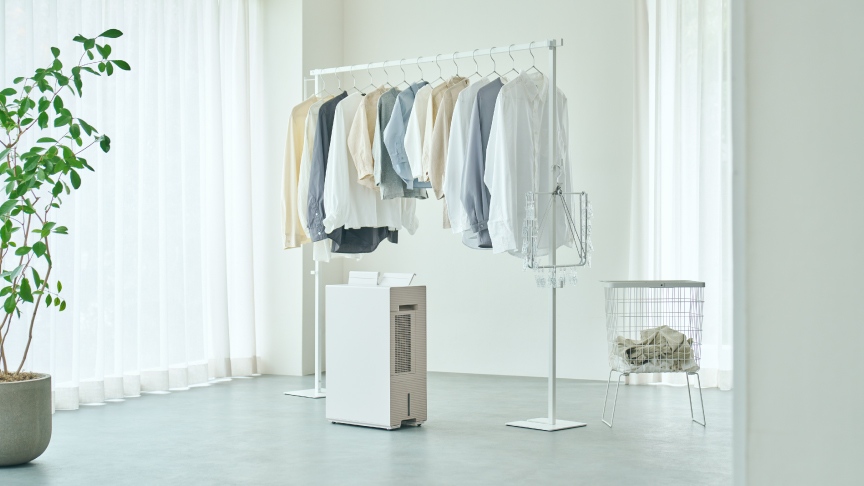 薄い色の服がかかったハンガーラックの下に、白い衣類乾燥除湿機が置かれている画像です。