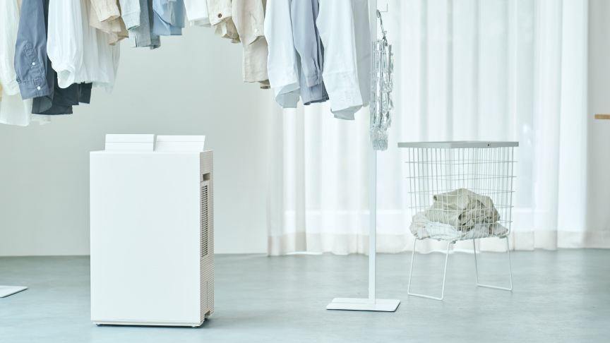 薄い色の服がかかったハンガーラックの下に、白い衣類乾燥除湿機が置かれており、その衣類乾燥除湿機をクローズアップした画像です。