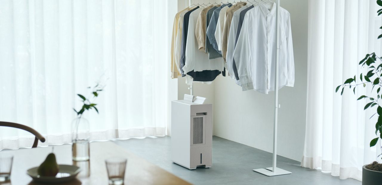薄い色の服がかかったハンガーラックの下に、白い衣類乾燥除湿機が置かれている画像です。