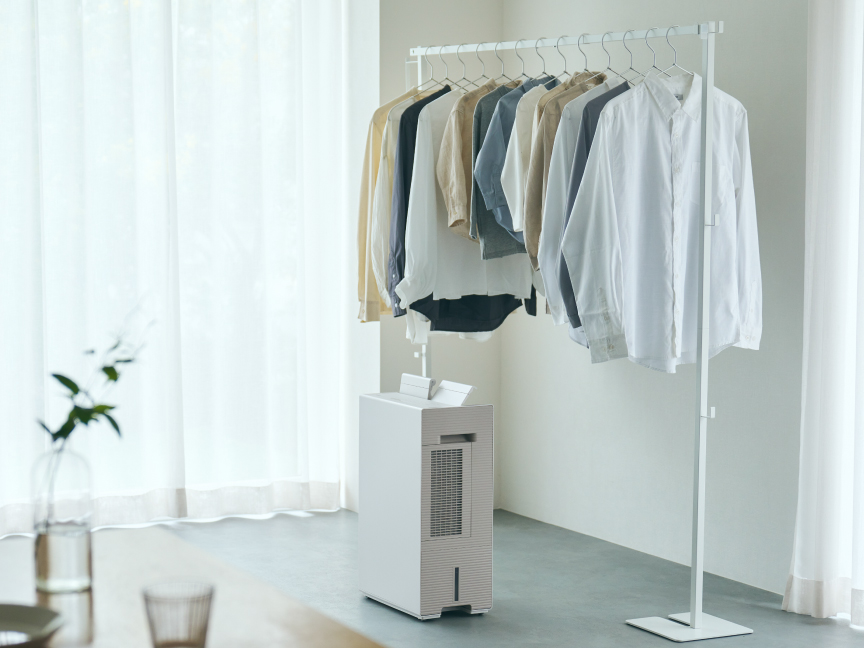 薄い色の服がかかったハンガーラックの下に、白い衣類乾燥除湿機が置かれている画像です。