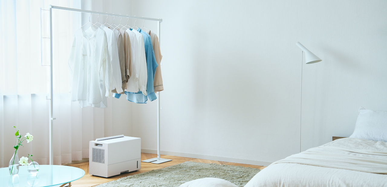 薄い色の服がかかったハンガーラックの下に、コンパクトな白い衣類乾燥除湿機が置かれている画像です。