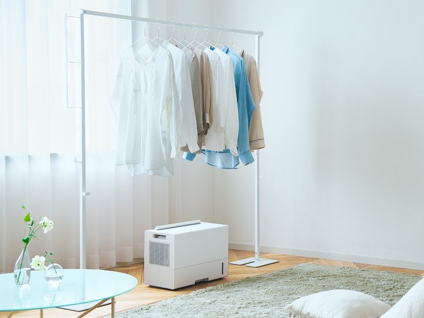 薄い色の服がかかったハンガーラックの下に、コンパクトな白い衣類乾燥除湿機が置かれている画像です。