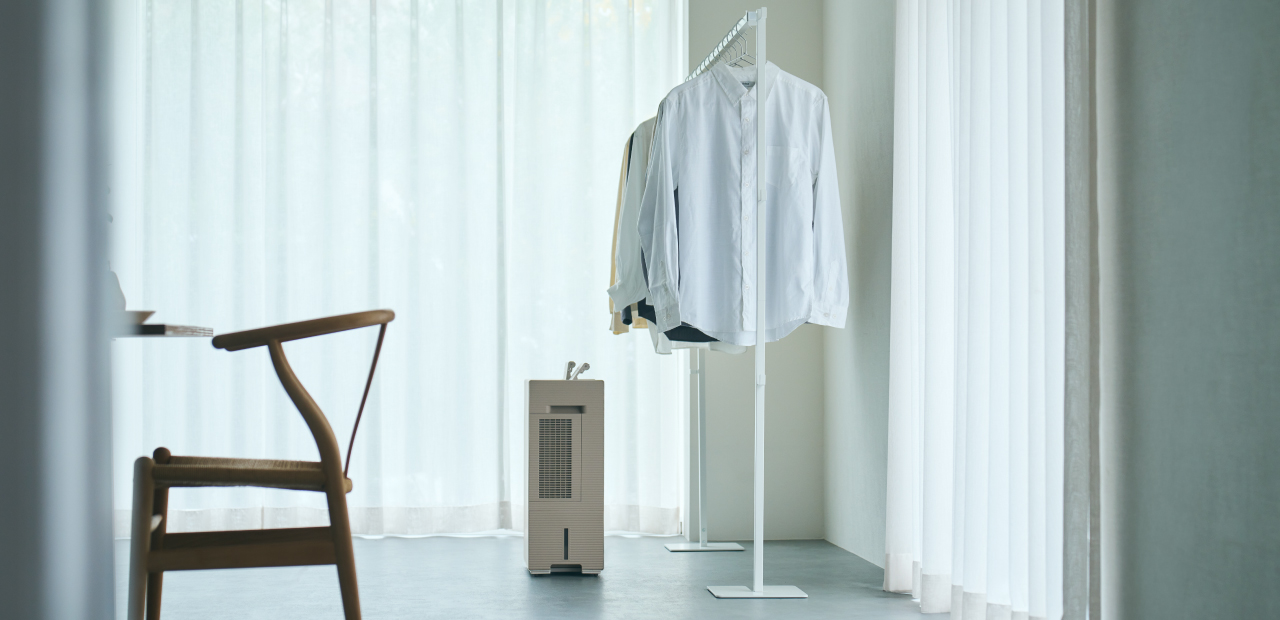 薄い色の服がかかったハンガーラックの下に、白い衣類乾燥除湿機が置かれている画像で、衣類乾燥除湿機の側面が見えています。