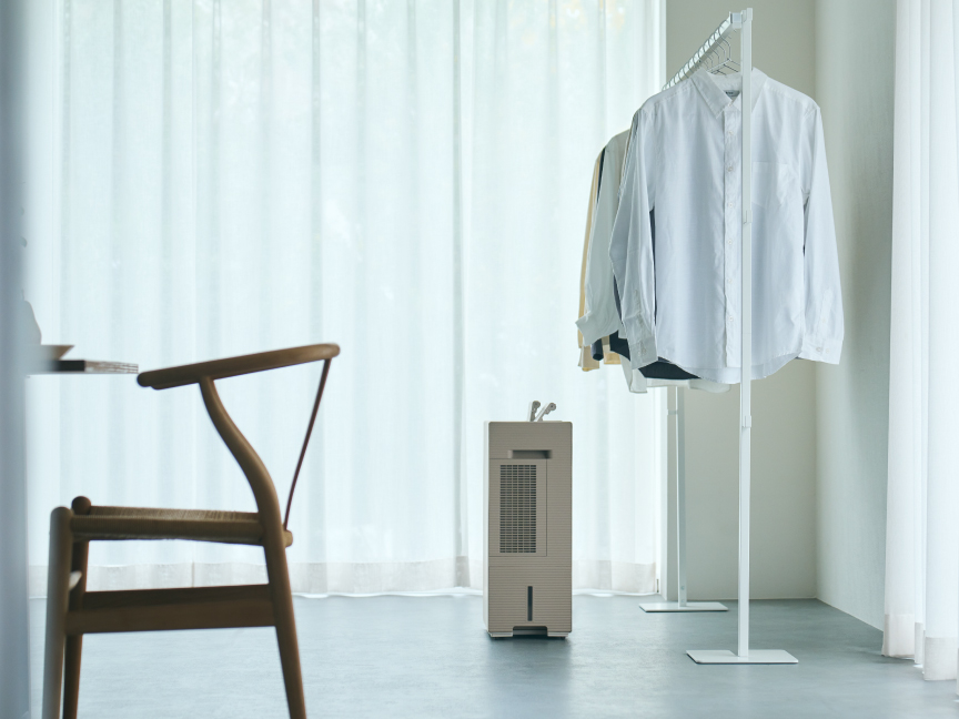 薄い色の服がかかったハンガーラックの下に、白い衣類乾燥除湿機が置かれている画像で、衣類乾燥除湿機の側面が見えています。