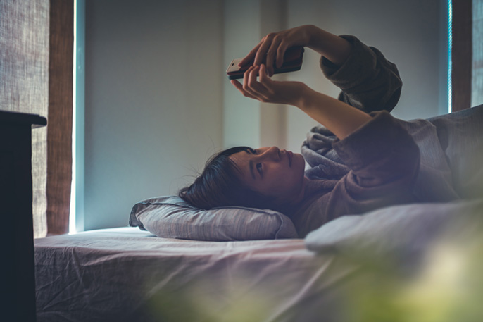 写真：ベッドに横になってスマホを使う女性
