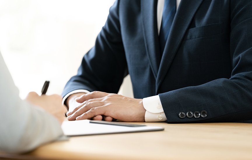 写真：打ち合わせや相談の場面のイメージ。スーツを着用した人物がデスクに着席し、手元にタブレットを置き、向かい側の相手と話をしている様子
