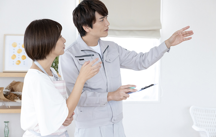 写真：住宅の相談・説明の場面のイメージ。作業着を着た説明者がクリップボードを手に室内の一方向を指し示し、同席者が説明を聞いている様子