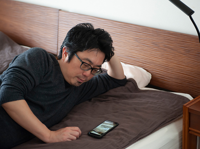 ベッドで横になりスマホでテレビを見ている男性の写真