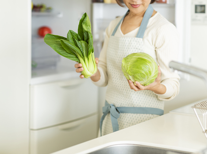 写真：冷蔵庫の前でキャベツとチンゲン菜を持った女性のイメージ