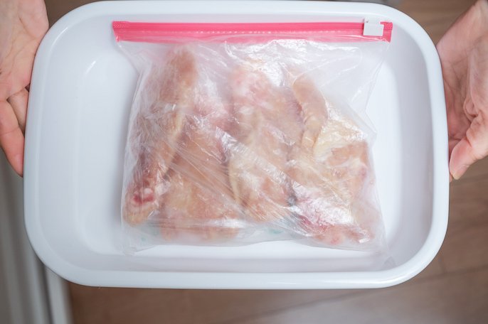 写真：ラップで包まれた肉が入っている冷凍用保存袋が、お皿の上に置かれ、それを両手で持っている様子