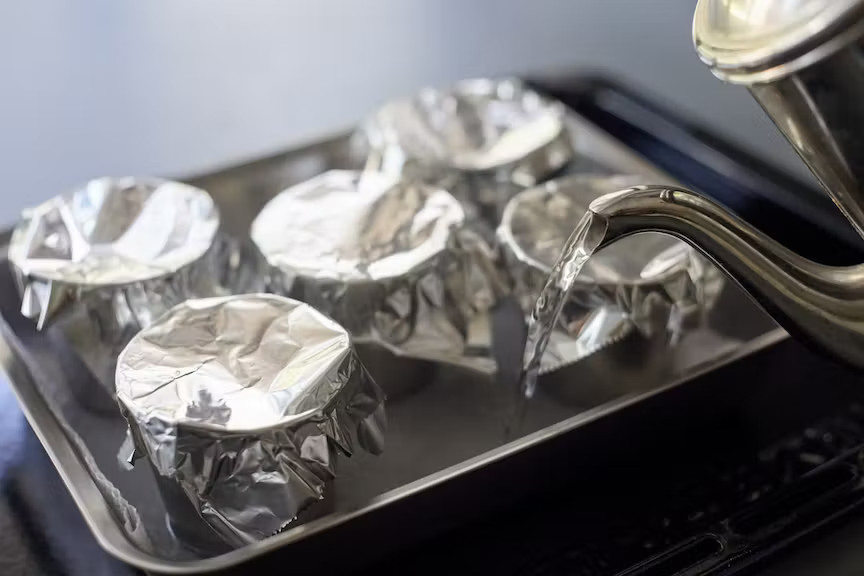 写真：キッチンペーパーを敷いたバットにプリンカップを並べ天板に置き、バットの中に50℃程度の湯を1cmの高さほど注ぐ様子