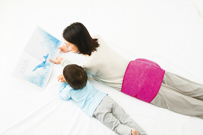 写真：うつ伏せで絵本の読み聞かせをしている母親と子ども。母親は腰にコードレス骨盤おしりリフレ（EW-RA79）を巻いている。