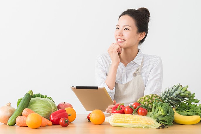 写真：女性がテーブル上に様々な野菜や果物を広げて見ながら思案しているイメージ
