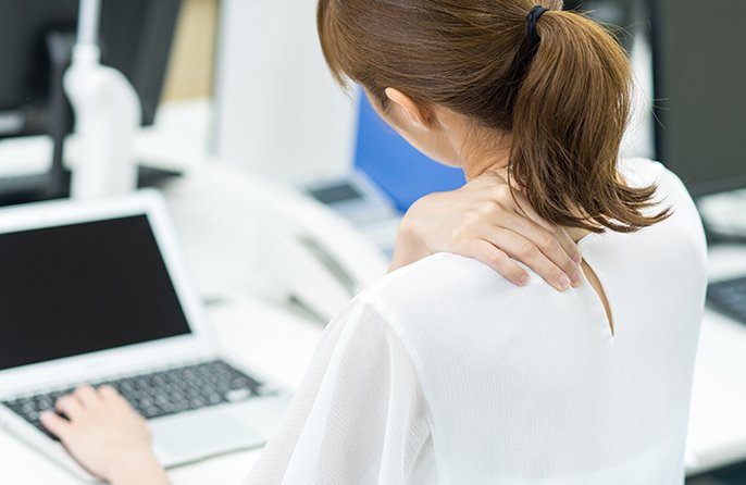 写真：パソコン作業をしている女性が、右手を左肩に当てて、首をかしげている様子