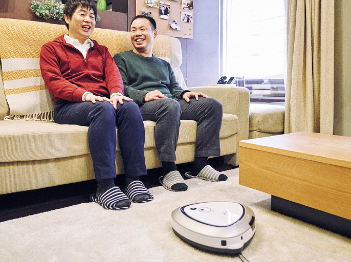 写真：ソファーに座って話をする二人と、床を走るロボット掃除機