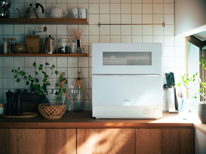 木製カウンターの上に白い食洗機が置かれた、観葉植物のある明るいキッチン。