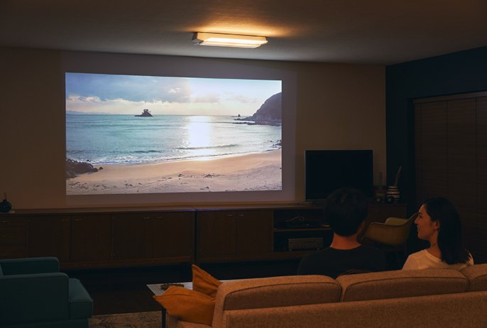 写真：スピーカー搭載シーリングライト【サウンドシリーズ】のある部屋にて、壁に投影された海岸の映像を見ながら、ソファーに座り会話している2人の様子