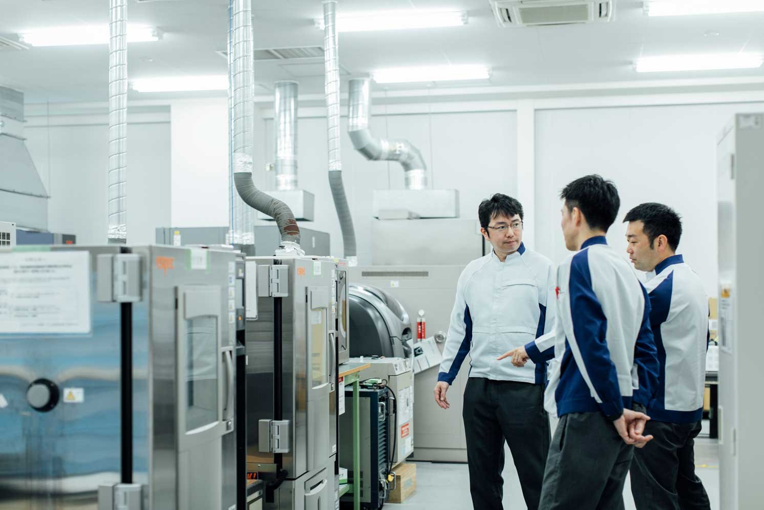 「再生プラスチックの寿命試験を行なう環境試験機」の前で話すプロジェクトメンバーの写真