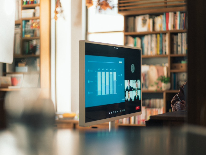 オンライン会議画面を表示したモニターが、書棚のある室内に置かれている。