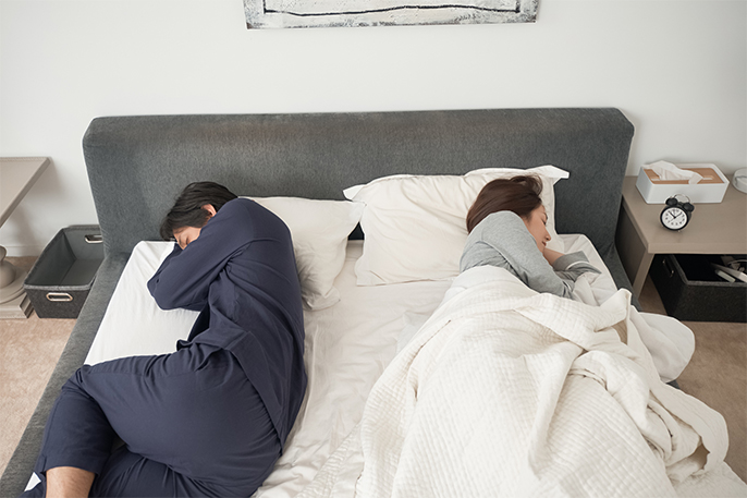 写真：ベッドでタオルケットにくるまって寝る妻と、何もかけずに寝る夫