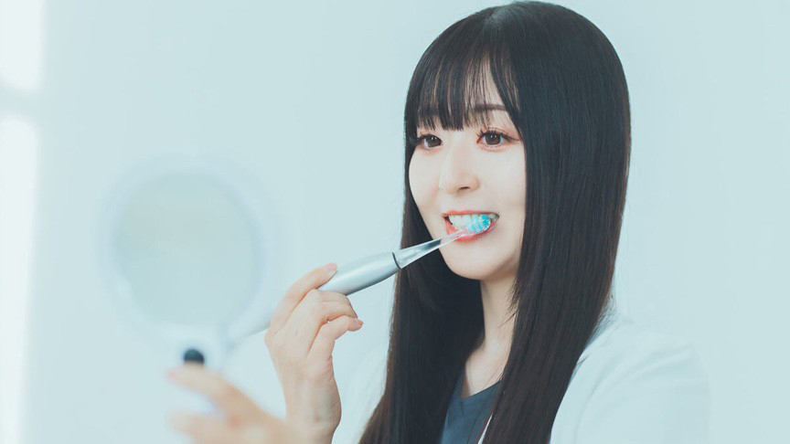 電動歯ブラシで歯を磨きながら手鏡を見ている女性